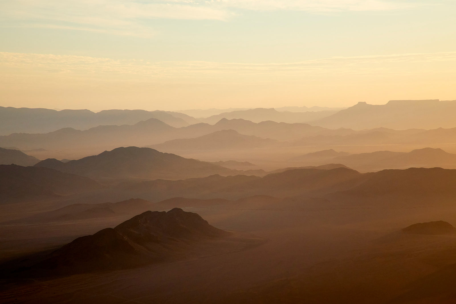 Aerial view of desert mountain ranges in soft golden light, layers of silhouetted ridges fading into mist