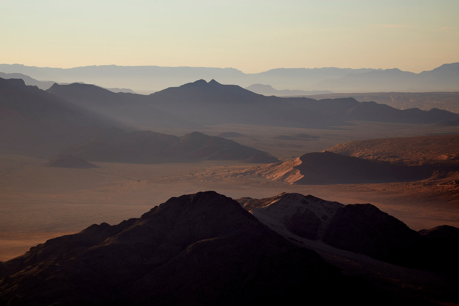Aerial view of rugged desert mountains with sun rays breaking through haze, dune visible in foreground