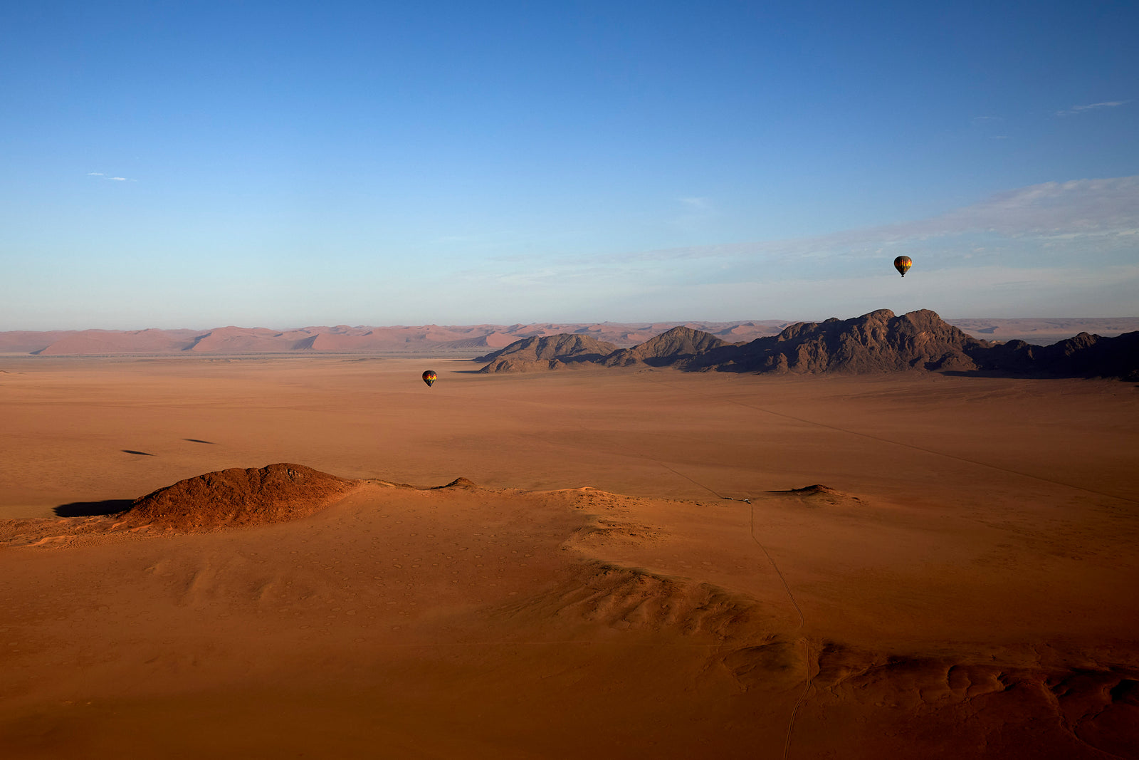 Two hot air balloons floating above vast Namibian desert plain with rocky outcrops and dunes at dawn