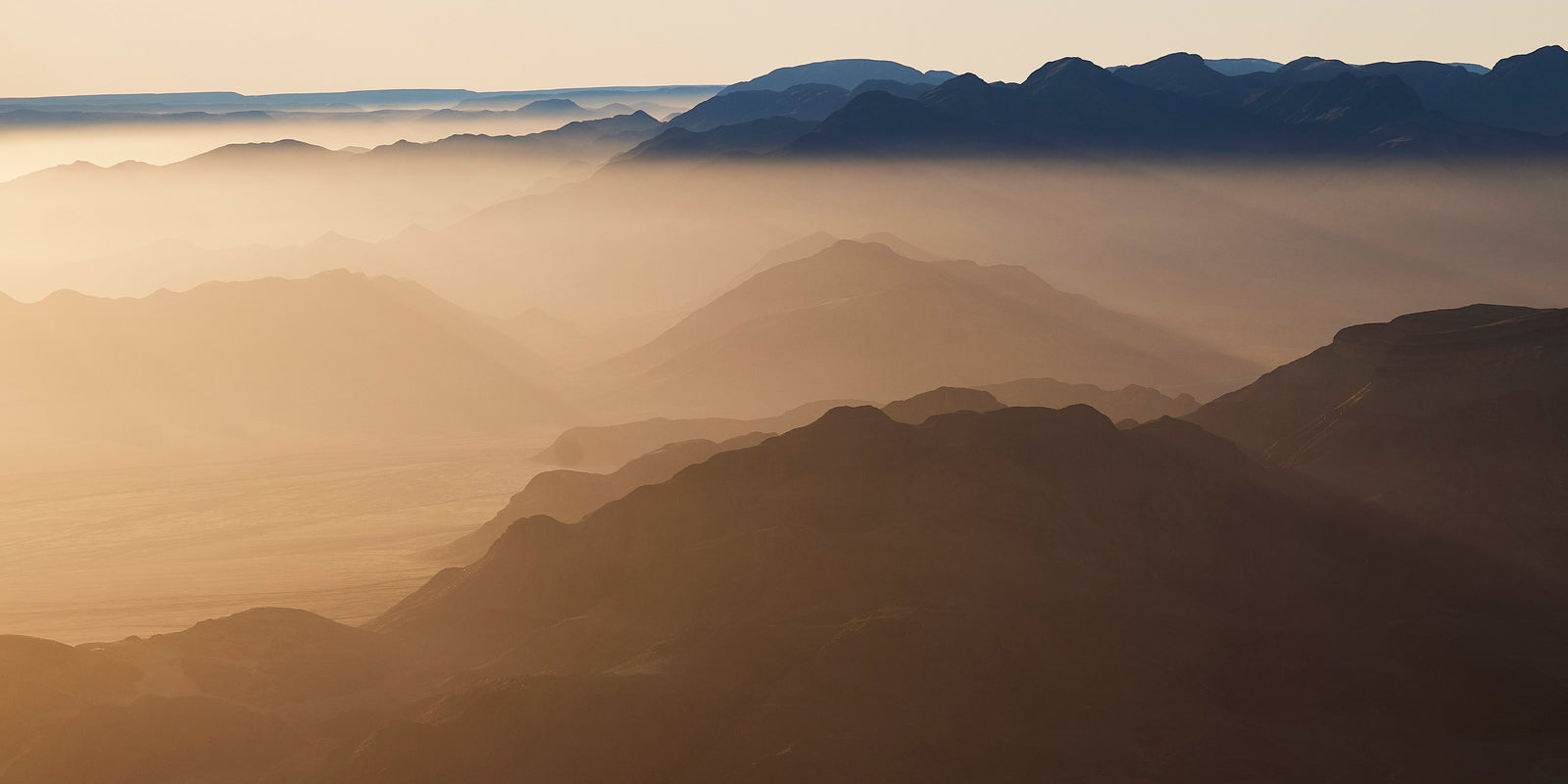 Panoramic aerial view of layered mountain ridges in golden haze, desert valleys stretching to the horizon