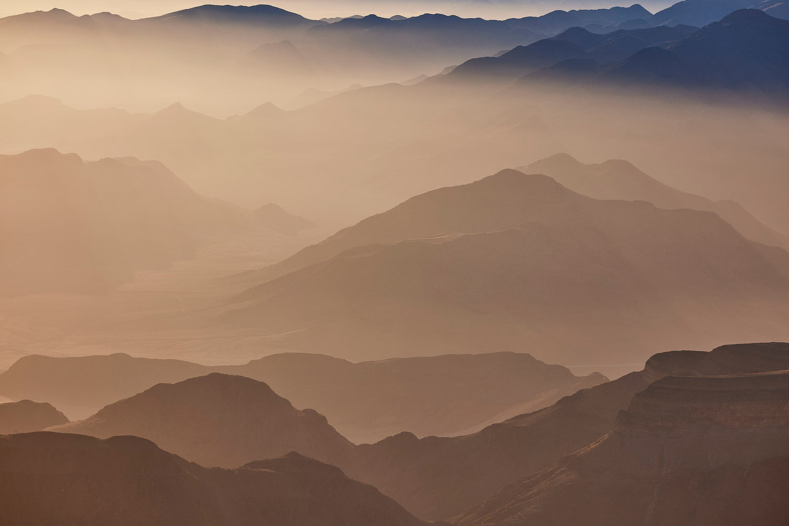 Aerial view of layered desert mountains fading into warm mist, soft tonal gradations from brown to blue