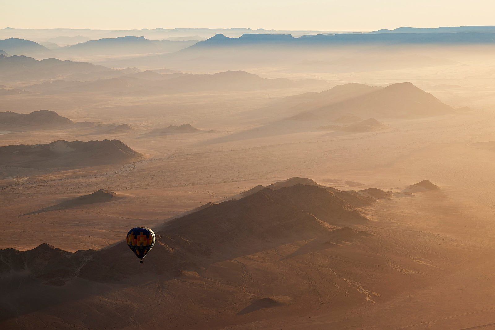 Hot air balloon floating low over hazy desert mountains at sunrise, golden light and layered ridges
