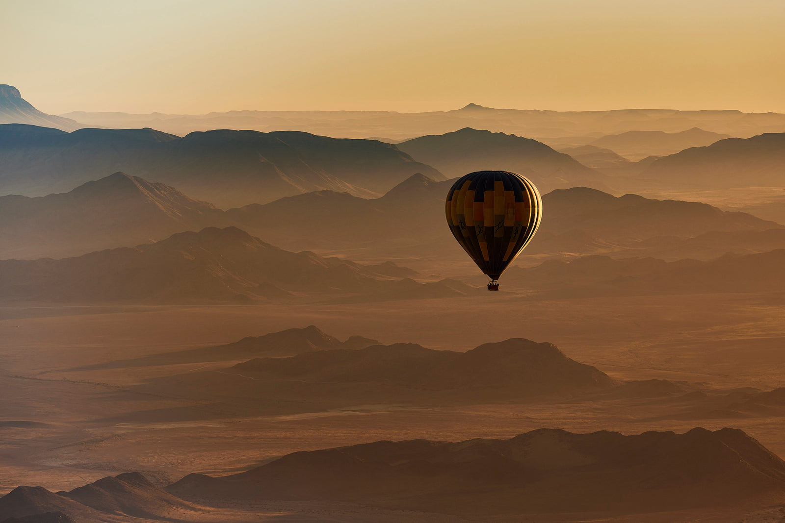 Hot air balloon silhouetted against golden haze over layered desert mountains at dawn