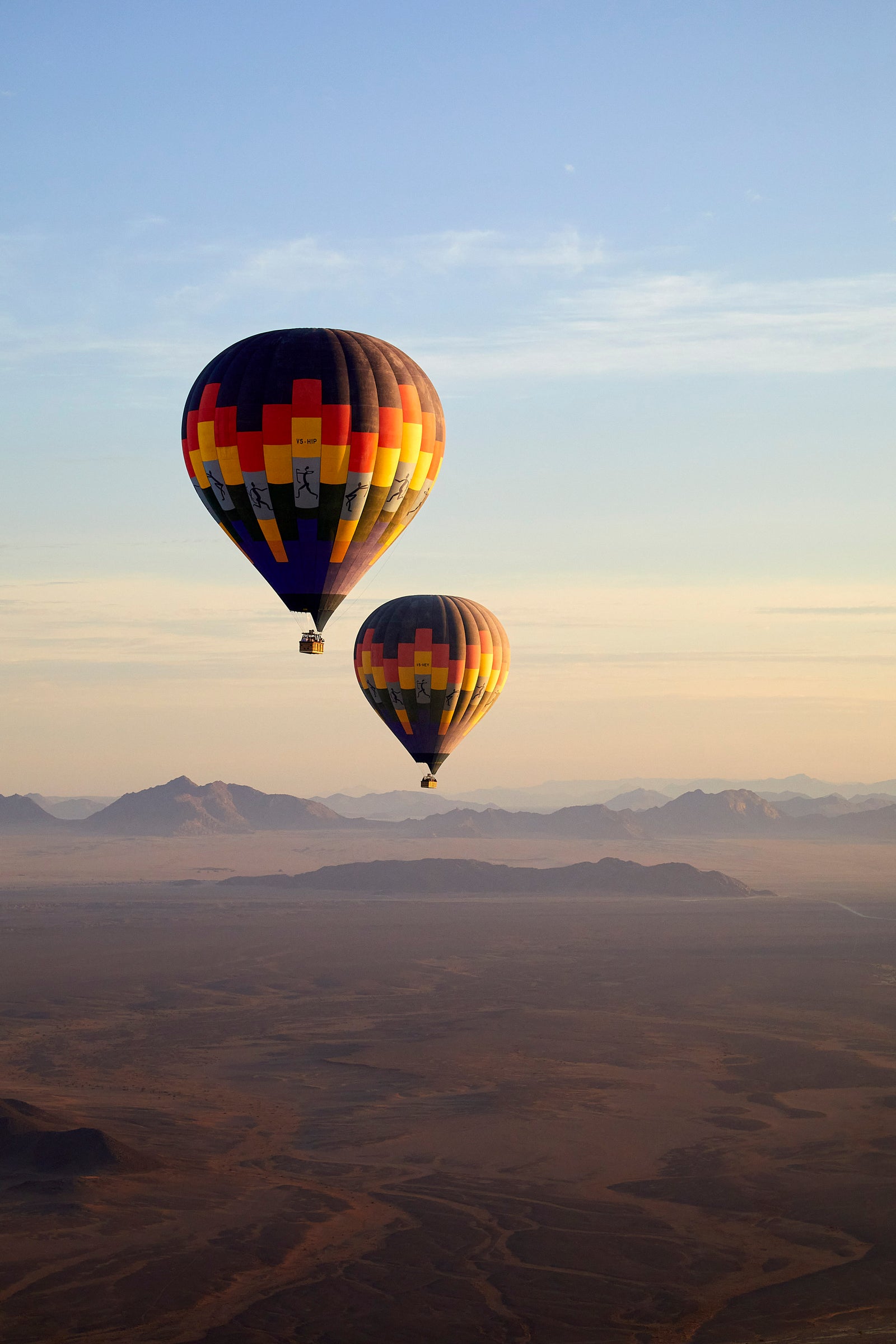 Two colourful hot air balloons ascending over misty desert plains with mountains in the distance