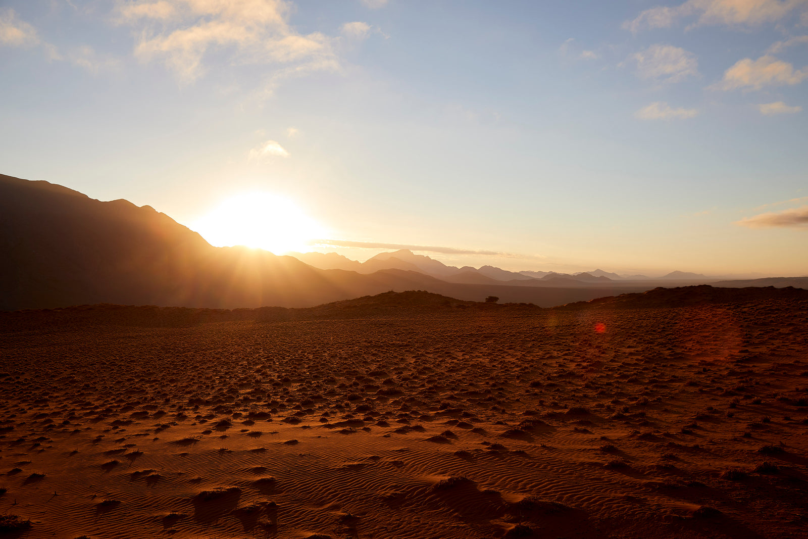 Sun setting behind desert mountains, starburst light over rippled sand plain 