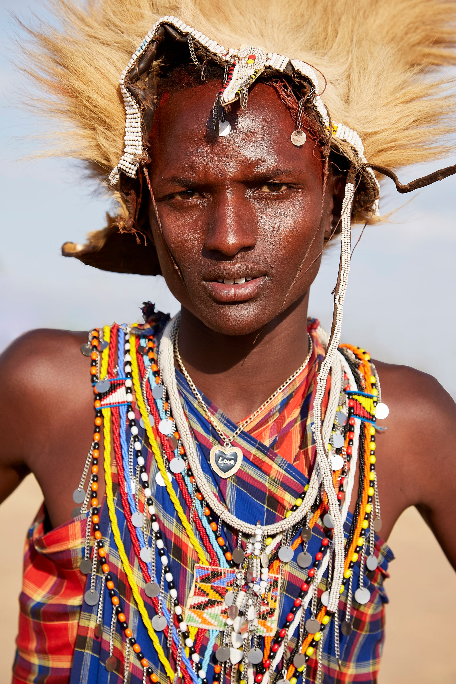 Portrait of a young Maasai warrior wearing a lion-mane headdress, beaded necklaces, and colourful shuka