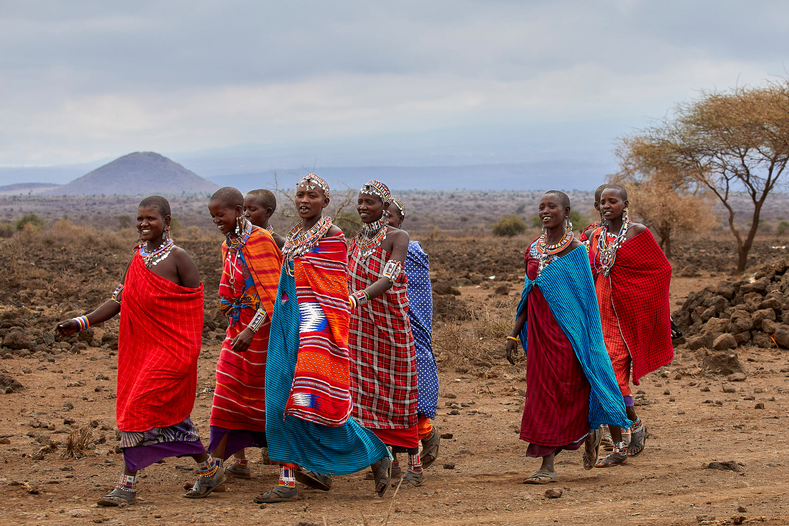 Group of Maasai women walking and laughing together in bright red, blue, and orange shukas, hill behind