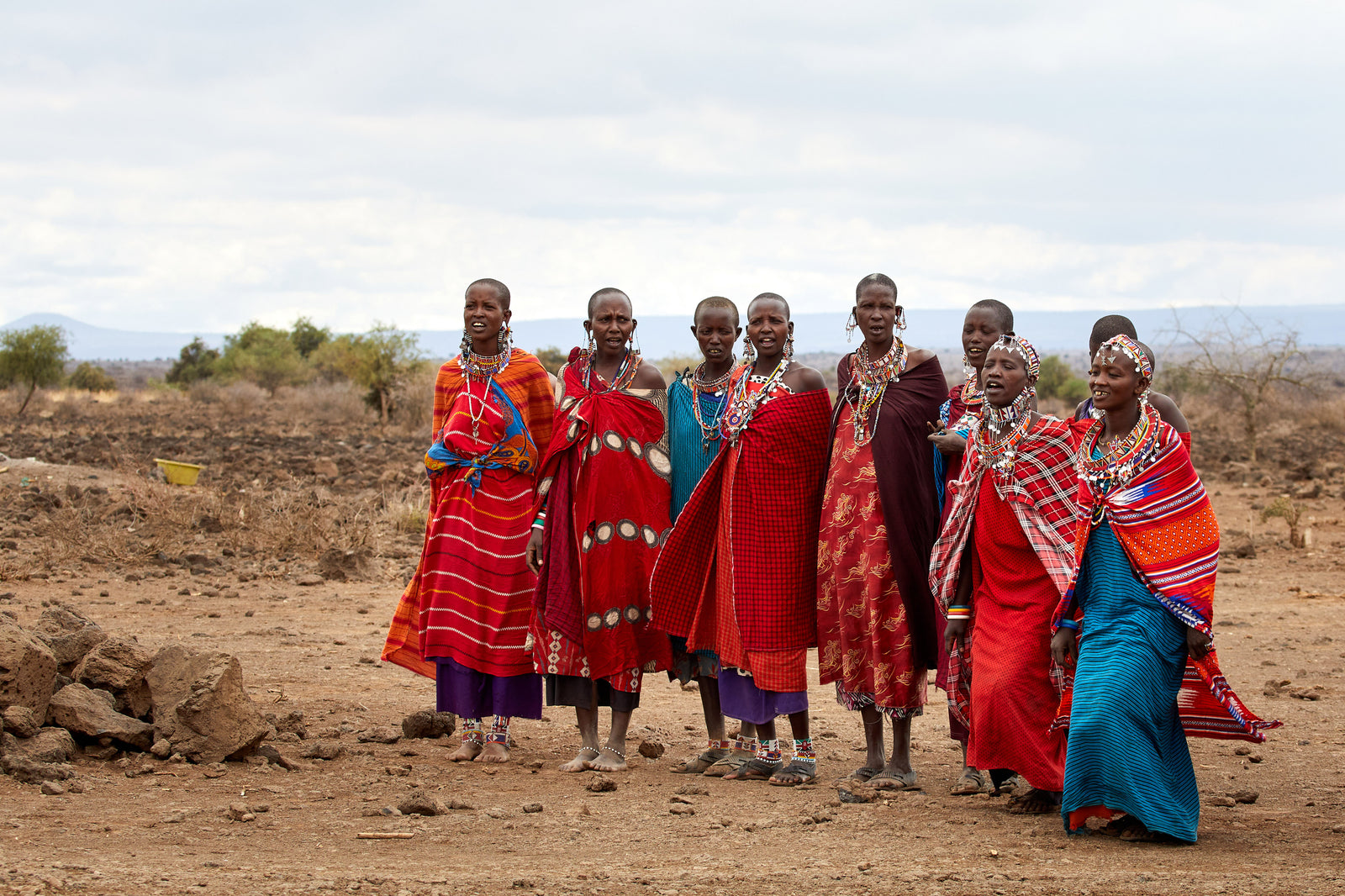 Group of Maasai women standing together in colourful wraps and beaded jewellery, dry landscape behind