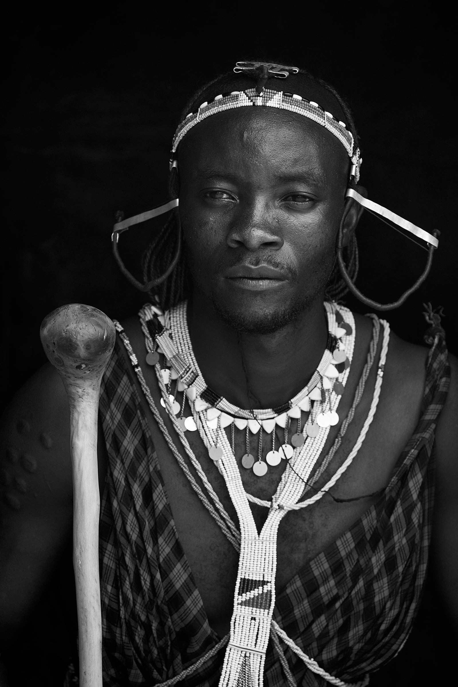 Black and white portrait of a Maasai man holding a wooden club, wearing beaded headband, earrings, and necklaces against a dark background