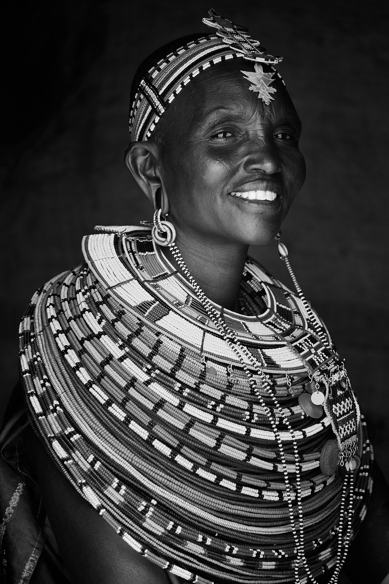 Black and white portrait of a Samburu woman smiling, wearing an elaborate layered beaded collar and headpiece