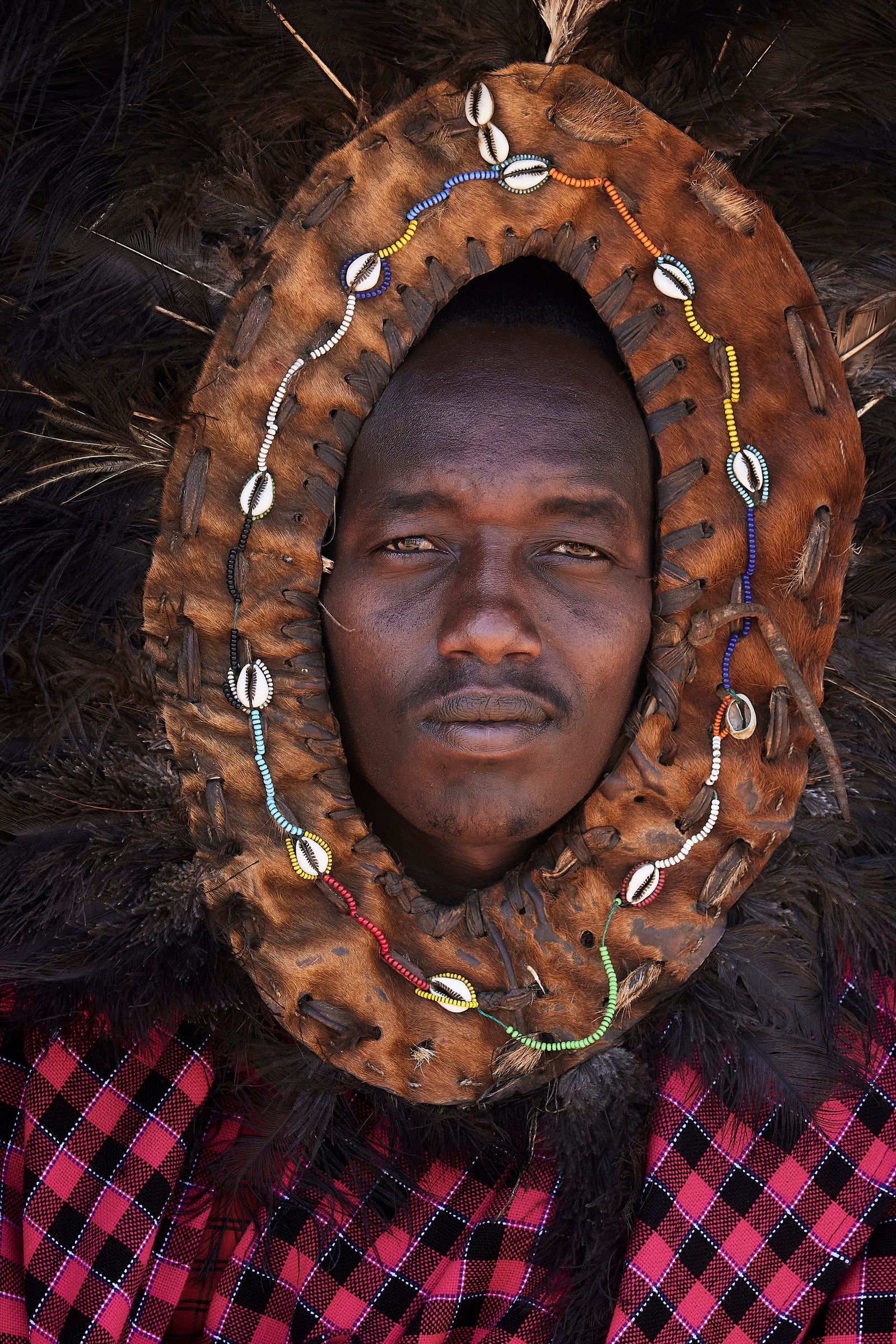 Portrait of a Maasai man wearing a ceremonial leather and cowrie-shell headdress framing his face, feather plume behind