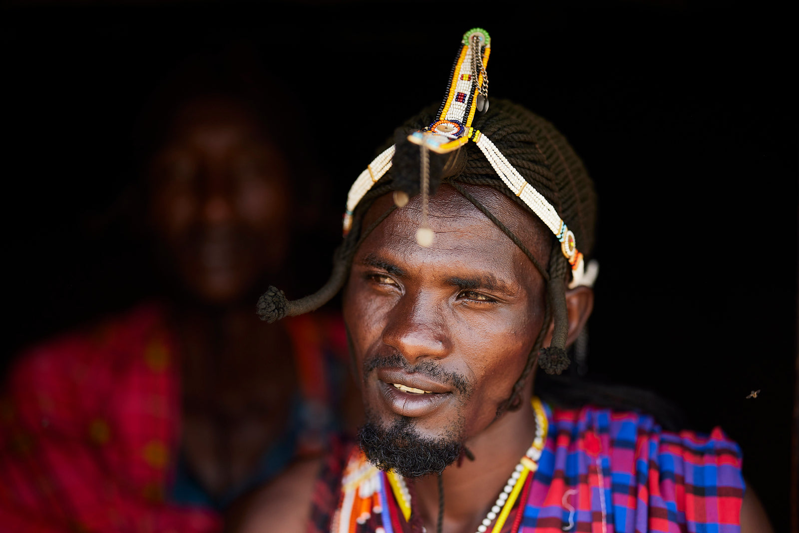 Portrait of a Maasai warrior with beaded headpiece and blue-red shuka, another figure softly visible behind