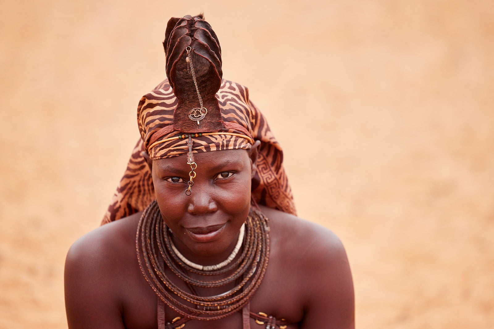 Portrait of a Himba woman looking at camera, wearing an animal-print headwrap, sculpted ochre hair crest, and beaded necklaces