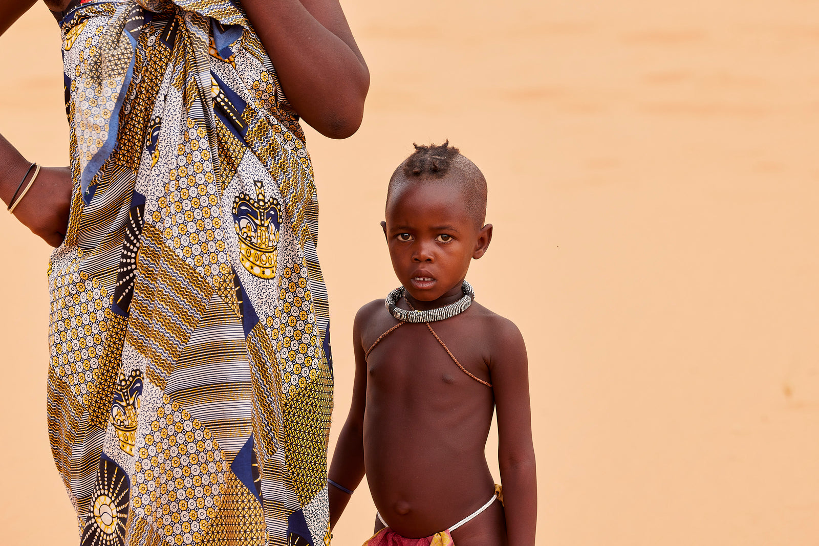 Young Himba child standing beside a woman's patterned wrap, wearing a simple beaded necklace, sandy background