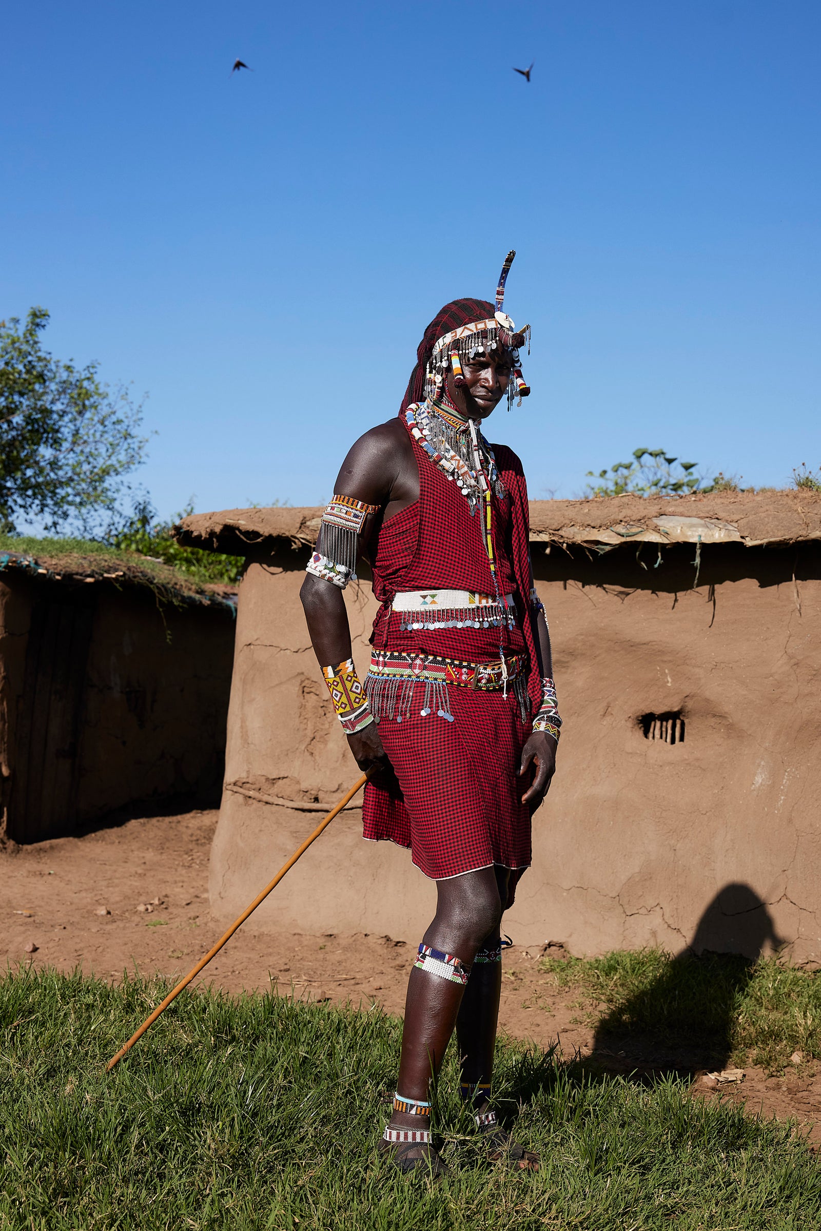 Full-length portrait of a Maasai warrior in ceremonial dress standing beside a mud hut, spear in hand, blue sky