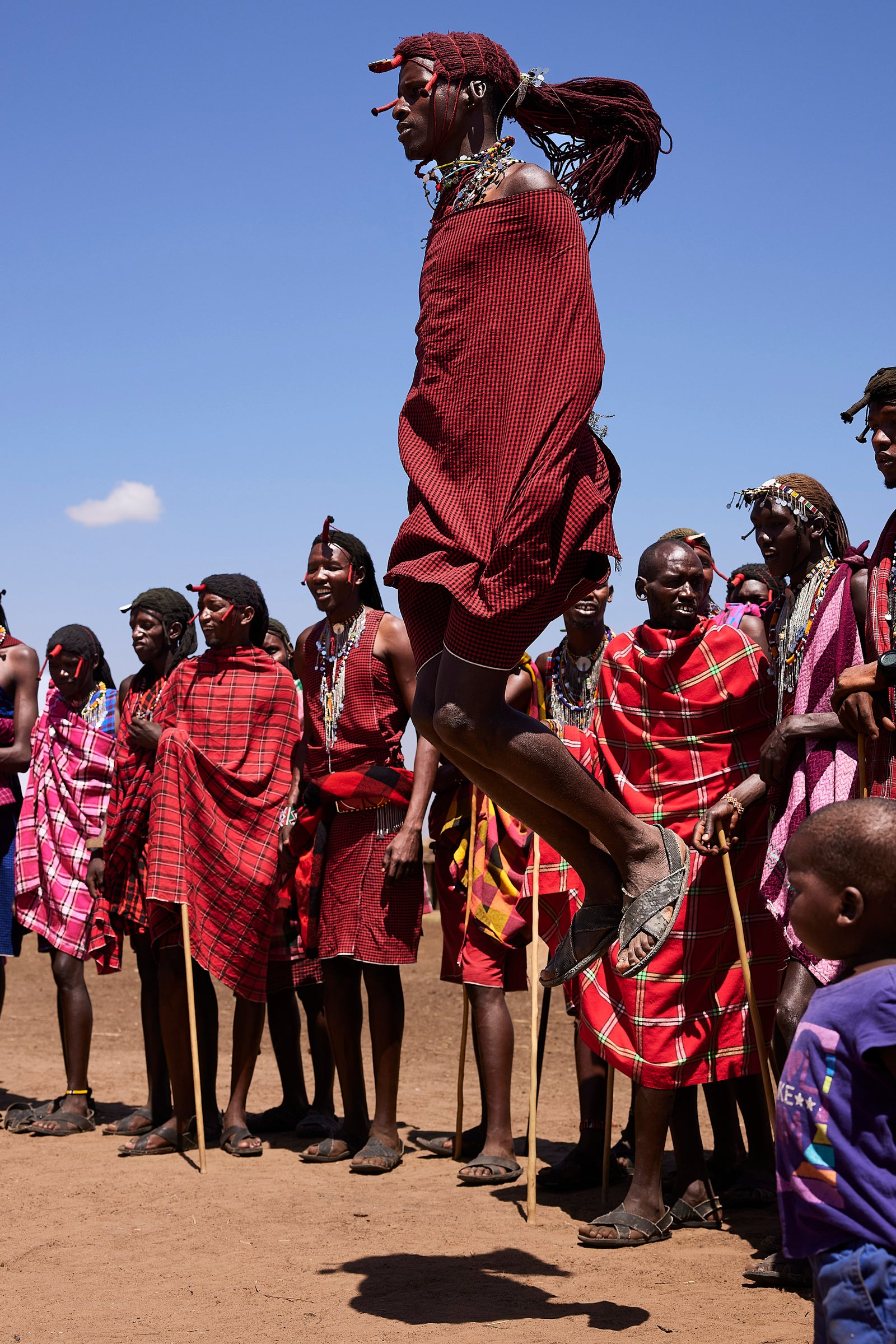 Maasai warrior leaping high in a traditional jumping dance, red shuka flowing, crowd watching behind